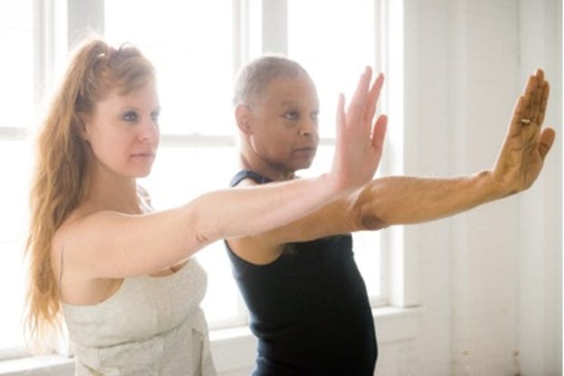 Close-up of two performers staring directly ahead and extending one arm forward with their palms facing up in front of drawn windows creating soft and hazy lighting. 