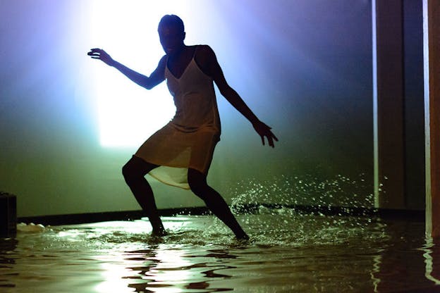 Okpokwasili with their mouth agape slightly crouching and bending one arm in front of them, standing in ankle-high water in a dimly-lit room streaked with green and purple light emanating from a bright white light in the background.