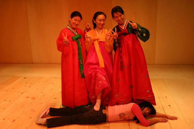 Three people dressed in traditional Korean wear make the peace sign with their hands and smile forward. The person in the middle steps on a woman dressed in modern wear as she lays on her stomach with her eyes closed and arms outstretched. The set has wood floors and walls.