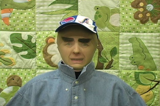 A video still of Ben Tor in front of a green quilt patterned with green animals and leaves. She appears bald and wears a blue baseball cap, blue button up shirt, and has very thick eyebrows. 