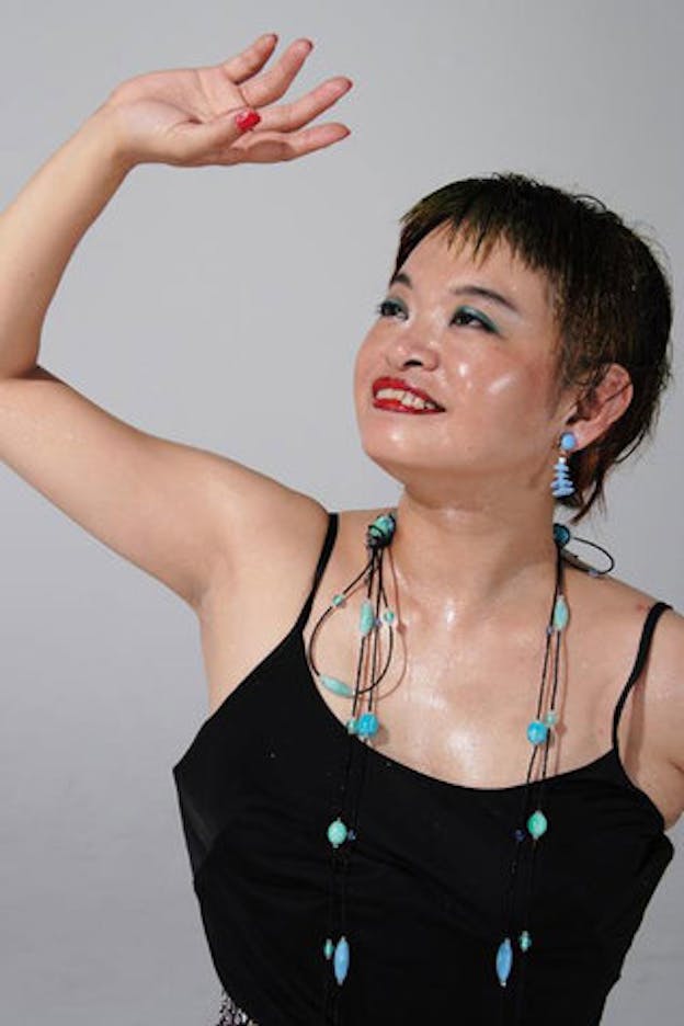 Nexus6 performs in a black tank top, blue beaded necklace, blue earrings, blue eyeshadow, and red lipstick. She raises her right hand upward and looks towards it, smiling. 