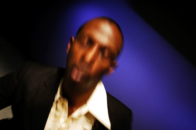  Blurred and slanted close-up of a person closing their eyes and wearing a tuxedo against an indigo and black background.