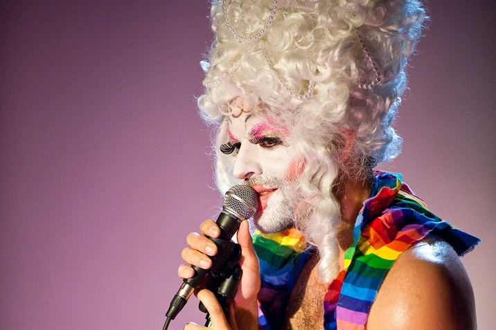 Side-profile of a performer wearing a curly pearl-strewn platinum beehive wig, thick white face paint, red blush, pink eyeshadow, and rainbow-colored cloth around their shoulders looks outwards and raises a microphone to their lips. 