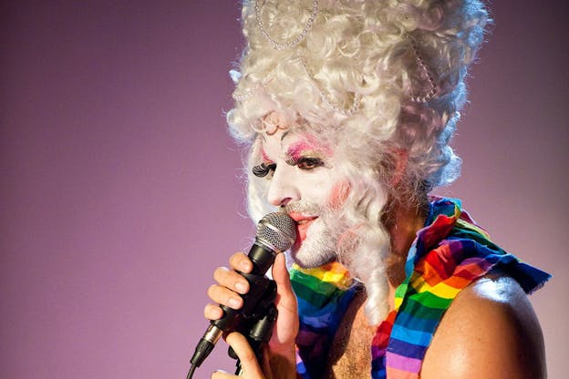 Side-profile of a performer wearing a curly pearl-strewn platinum beehive wig, thick white face paint, red blush, pink eyeshadow, and rainbow-colored cloth around their shoulders looks outwards and raises a microphone to their lips. 