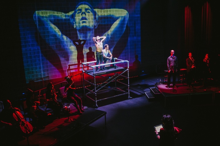 Two people on an elevated platform in a red and blue lighted room. One of them sits as the other stands shirtless, with the arms lifted and the hands on either side of the head copying the projection behind them on the stage. On either side of the platform musicians hidden by shadows and red lights.