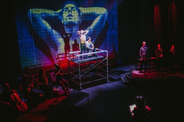 Two people on an elevated platform in a red and blue lighted room. One of them sits as the other stands shirtless, with the arms lifted and the hands on either side of the head copying the projection behind them on the stage. On either side of the platform musicians hidden by shadows and red lights.