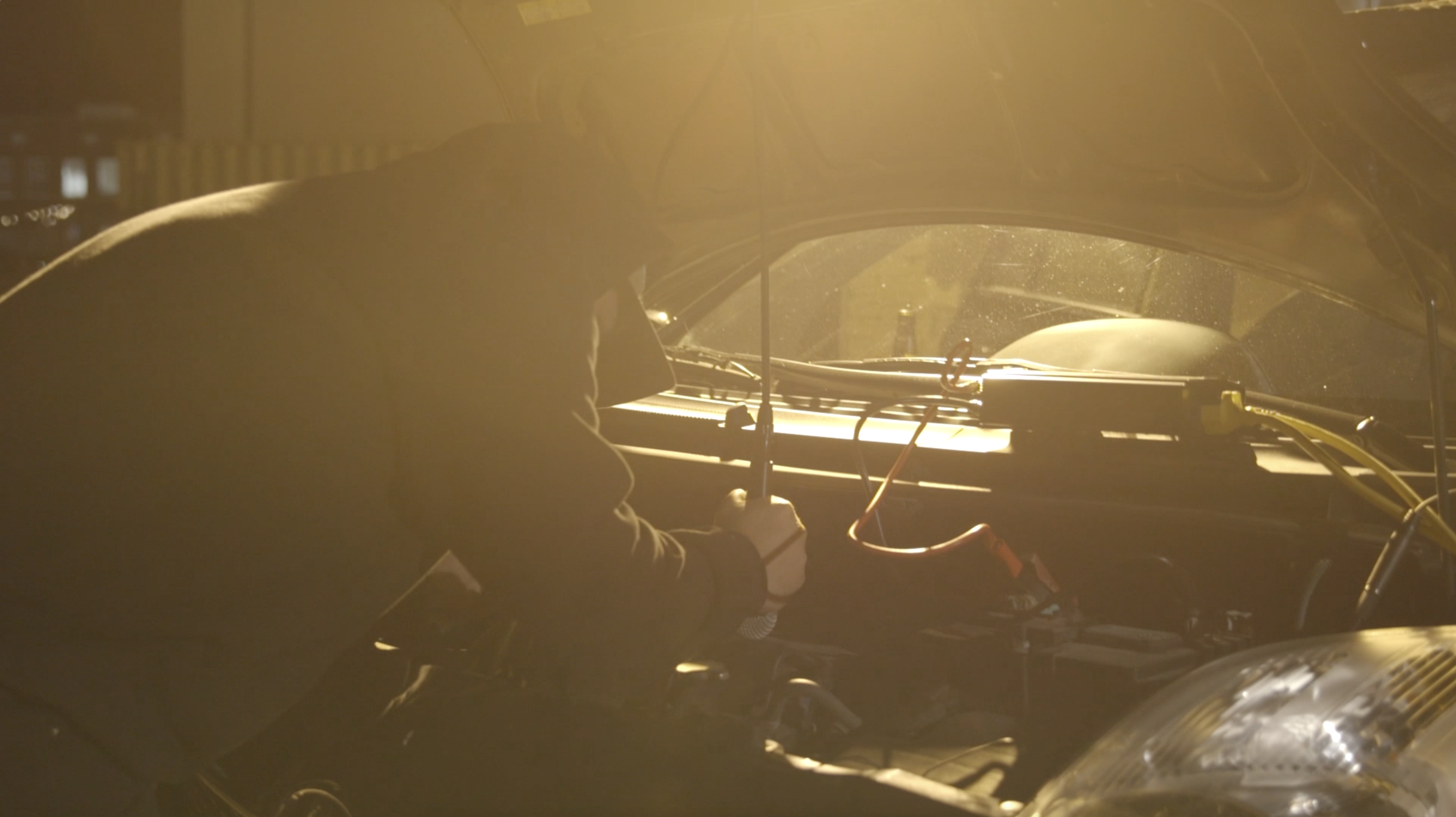 On the left side of the frame, Nathan Young is visible from the hips up and wears a dark hoodie and a baseball cap. He leans over the open hood of a car with his back to the camera, washed in yellow light. He holds a microphone vertically in his right hand, pointed downward to capture sound emitted by the car's engine.