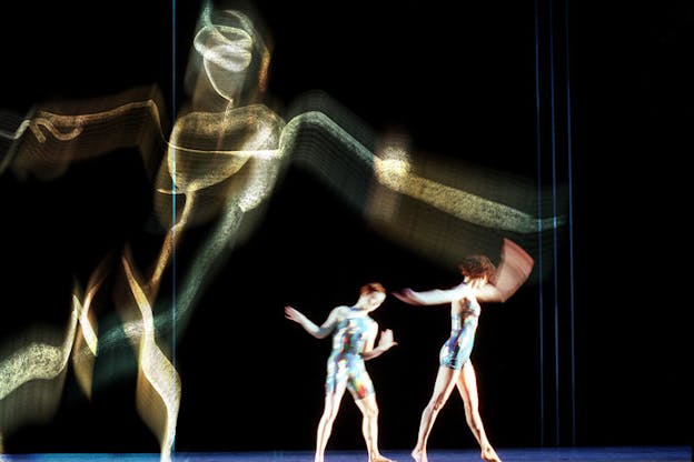 Two performers stand in the center of a dark stage wearing leotards with a metallic blue toned abstract pattern on them. Both peformers face the right side and step towards it, lifting their arms by their sides. A large gold sketch of a human figure is projected onto the otherwise black wall behind them. This figure leans towards the right. 