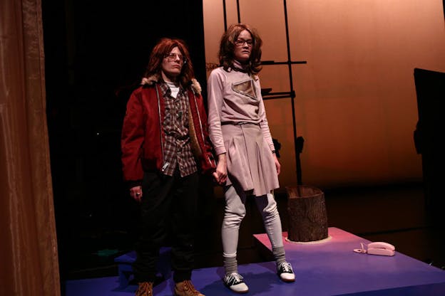 Two performers wearing brown wigs stand next to each other on a stage. The one on the left is dressed in a a red jacket, a tartan button up and dark jeans and holds the hand of the other performer dressed in a pink shirt and skirt.  