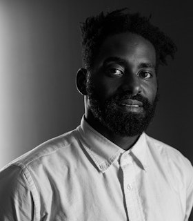 Photograph of Jerron Herman against a grey background, dressed in a white button up shirt, facing the camera, smiling slightly.