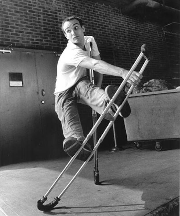 Bill Shannon performs in a space with brick walls. He holds his body off the ground by supporting his weight on a crutch under his left arm. He extends his legs out long in front of him and pushes them against another crutch which is diagonal to the floor and held up by his other arm. 