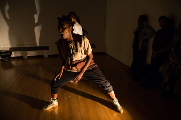 A performer lunges diagonally with their mouth open, suggesting speech. They wear a tan t-shirt, black sweatpants and white sneakers. A warm light floods the space from a position on the floor, casting shadows onto other performers who stand against a white wall in the background. 