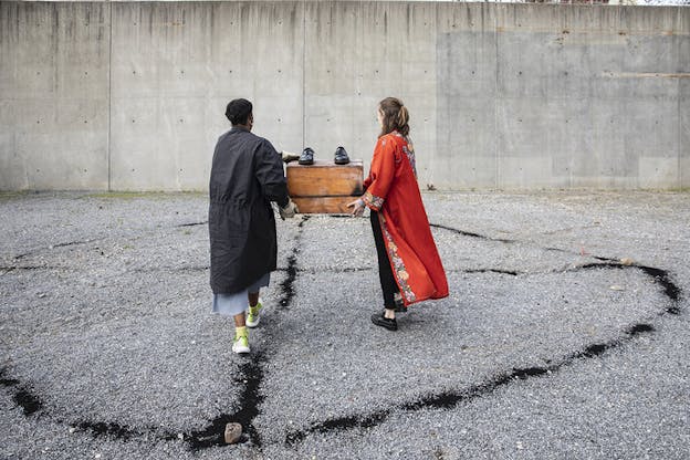 Two performers hold a wooden box between them and face away from viewers. Ontop of the wooden box, there are two dress shoes. One performer wears a red robe and the other wears a black robe. The performers walk on light grey gravel that is painted with a black circle pattern. 