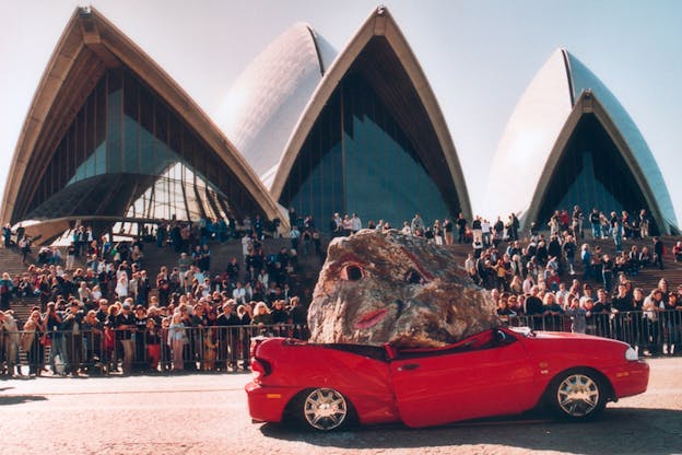 An audience in front of a building with triangular shapes watches a boulder with a face painted it on it crashing down on the roof of a red car.
