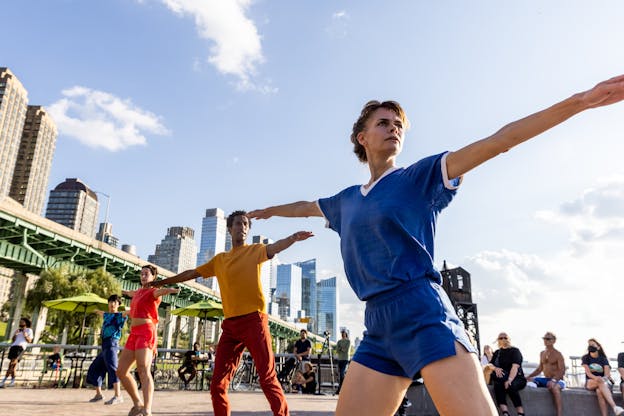 Four dancers perform outdoors in the sun on the Hudson River waterfront. Manhattan's skyscrapers are visible in the background along with a few audience members. The four dancers stand in a line with Joanna Kotze closest to the camera on the right side of the frame. The other three dancers stand behind her diagonally, with the fourth dancer at a distance from the camera on the left side of the frame. They all hold the same pose; their bodies are angled towards the left and their heads are turned over their left shoulders to look towards the right. They stand with their feet apart with their right knees bent, their arms are stretched out straight on either side. They all wear brightly colored clothing, some in shorts, some in pants, with t-shirts.
