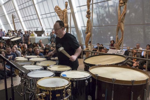 Winant plays an arrangement of varying drums. Behind them, an audience sits and watches. 