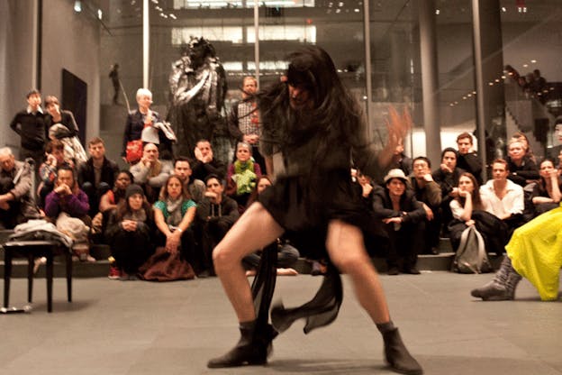 Performer in loose black garb thrashes their body, shaking their long hair in front of a small audience in a museum setting.
