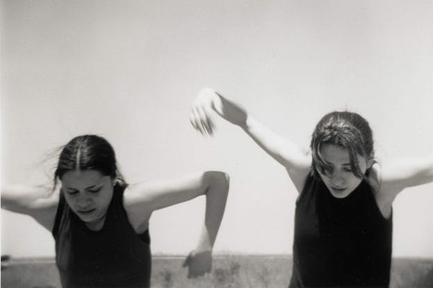 A black and white performance still of two performers wearing dark colored tank tops. The performer on the left looks downwards with their arms raised at ninety degree angles above their head. The performer on the right also looks down with their arms unbent and raised by their head. In the background there is a clear sky and a grassy field.