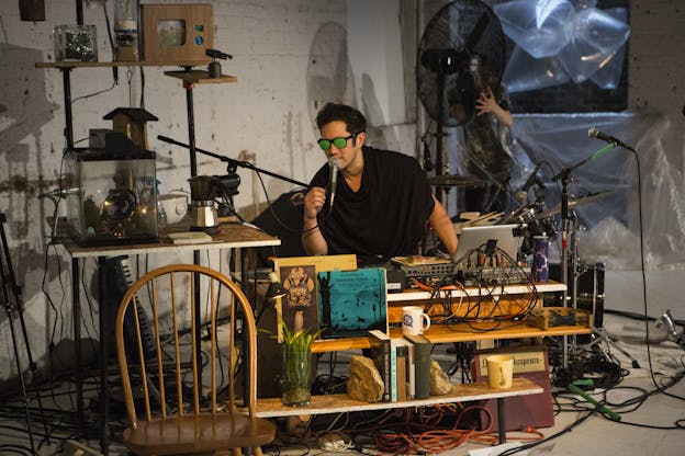 Tei Blow stands behind two tables covered with books and equipment in a softly lit room with white painted brick walls. He is bent forward as he speaks into a microphone. He is wearing a black shortsleeved wrapped tunic and mirrored sunglasses over his eyes.