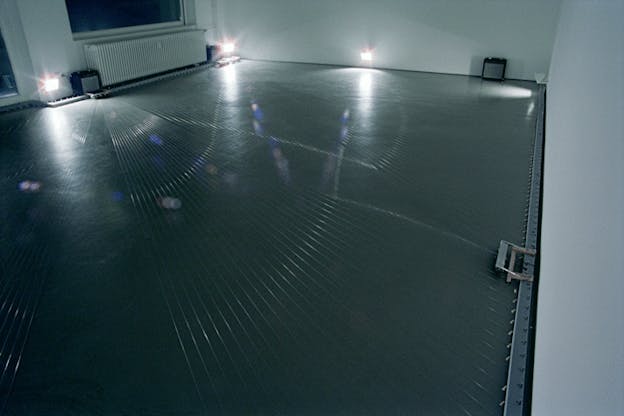 An image of a grey room with numerous silver strings stretched from wall to wall on the floor and two small amplifiers against the wall. Light from three fluorescent lamps refracts across the strings.   
