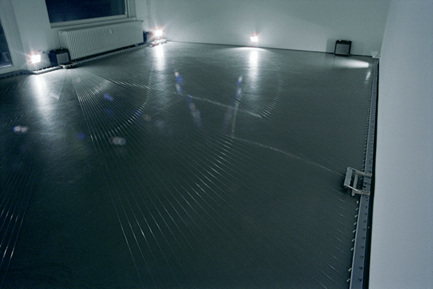 An image of a grey room with numerous silver strings stretched from wall to wall on the floor and two small amplifiers against the wall. Light from three fluorescent lamps refracts across the strings.   