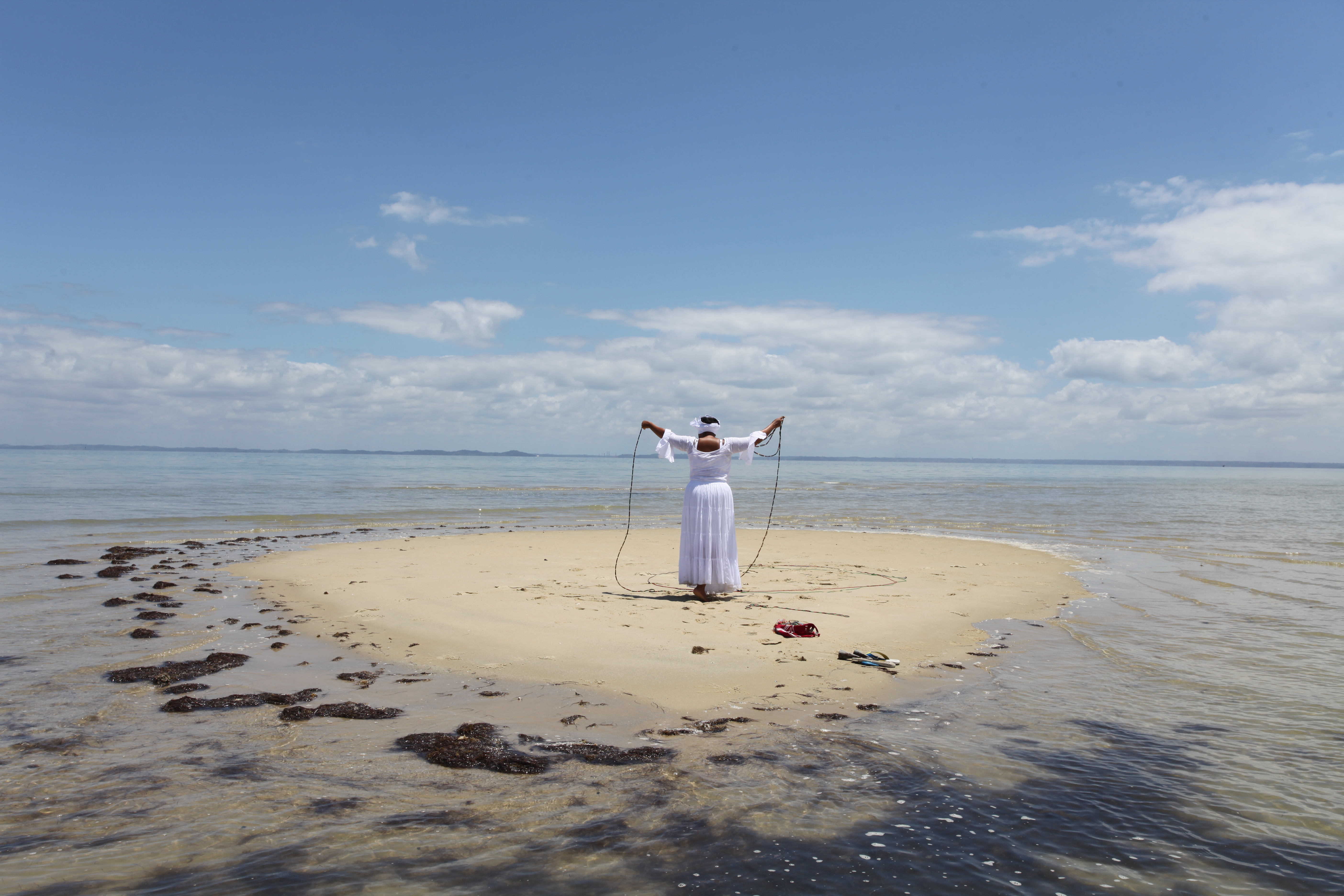 A figure clad in a long flowy white dress and headpiece stands with their back to the viewer on a small sand island surrounded by water. Their hands are extended upwards holding a black rope.