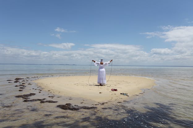 A figure clad in a long flowy white dress and headpiece stands with their back to the viewer on a small sand island surrounded by water. Their hands are extended upwards holding a black rope.