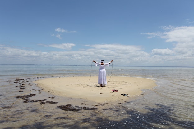 A figure clad in a long flowy white dress and headpiece stands with their back to the viewer on a small sand island surrounded by water. Their hands are extended upwards holding a black rope.
