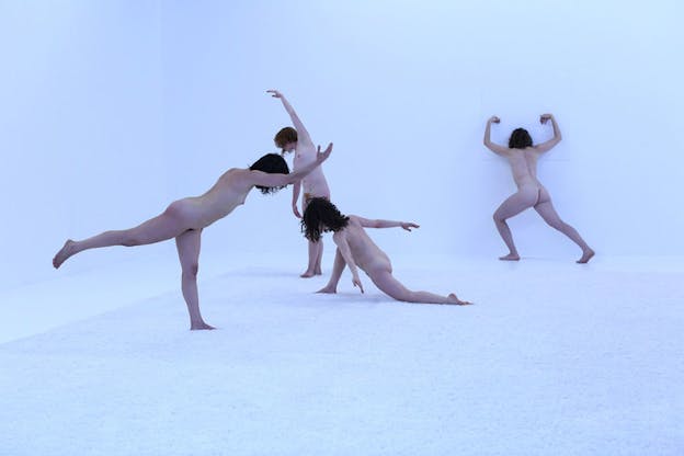 Four naked performers in various stances. The one closest to the viewer has one leg lifted backward with one hand forwards. The performer behind them kneels to the ground. Behind them the third performer stands with one arm raised. And in the back furthest from the viewer the fourth performer stands facing the wall flexing their biceps. 