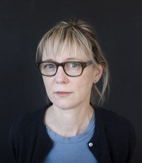 A portrait of Suzanne Bocanegra dressed in a blue shirt and black cardigan. Her blonde hair bangs touch her glasses