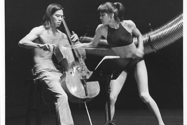 A black and white photograph of two performers in front of a long and curved aluminum duct hung from above. The performer on the left wearing a pair of pants plays the cello with a bow while looking down at the music stand. The performer on the right wearing a bralette and a brief underwear attempts to pull the cello bow out of the other performer's hand. The stage has a light color floor and a dark backdrop.