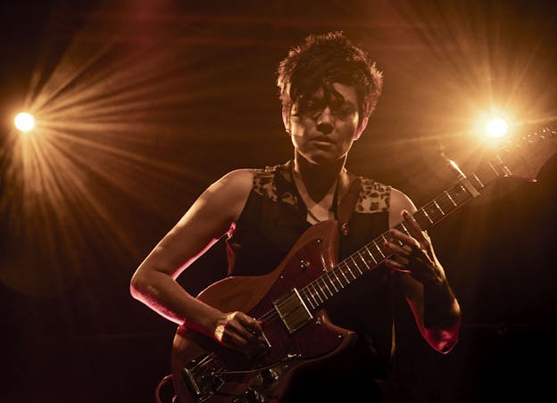 Ava Mendoza plays a wood grain patterned electric guitar on a dark stage, lit from behind by two bright orange lights. Visible from the hips up, their body and head are facing the camera, and their eyes gaze down at the floor. Their left hand holds the neck of the guitar while their right picks at the strings over the guitar body. They are wearing a black sleeveless button up top with  leopard print highlights on the shoulders, a chain necklace, and an earring in their right earlobe.