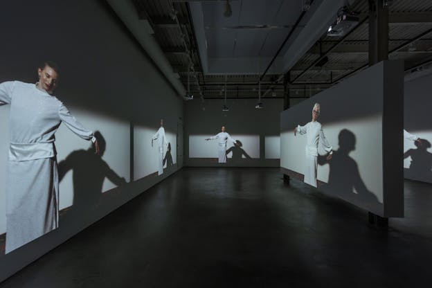 Installation view of figures clad in white with their arms open wide projected on the walls of a makeshift hallway. 