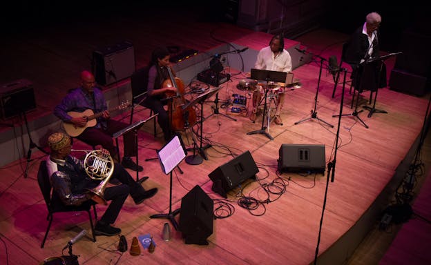 An overhead view of performers on a stage, surrounded by sheet music, amps, and microphones. Newman Taylor Baker sits on the right with his washboard and cymbals next to his collaborators, who are playing the cello, acoustic guitar, and French horn.
