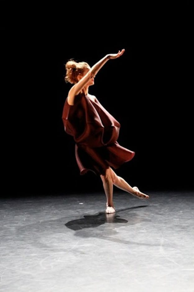 Performer wearing a flowy wine colored dress sweeps one leg behind them and curves one arm upwards, creating a crescent shape with their body on a gray stage with an ink black backdrop. 