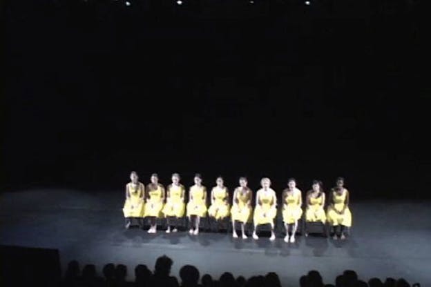 Several performers wearing yellow dresses sit besides each other in a dark space.