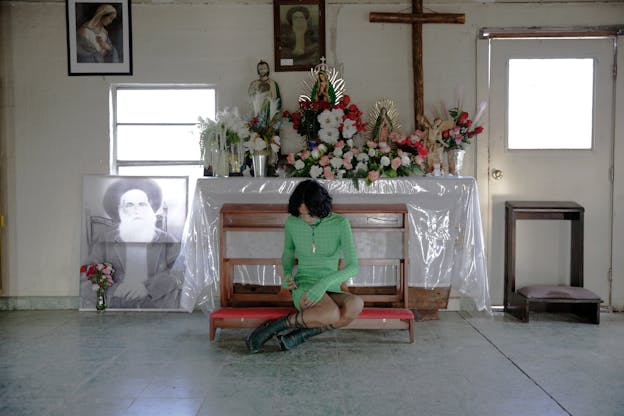 Angel Lartigue sits on a kneeler in front of an altar with flowers, a cross, and various religious objects including two statues of the Virgin of Guadalupe. She is wearing a green dress, a necklace, and is injecting estrogen into her thigh with a medical needle.