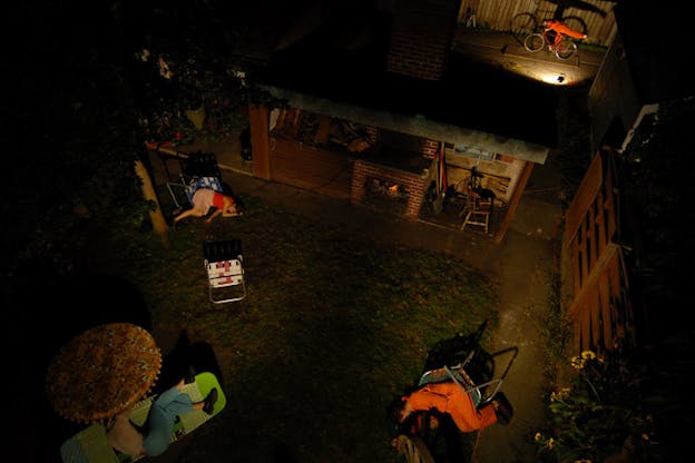 Aerial night view of three reposing bodies leaning against tipped lawn chairs in a dimly lit backyard.