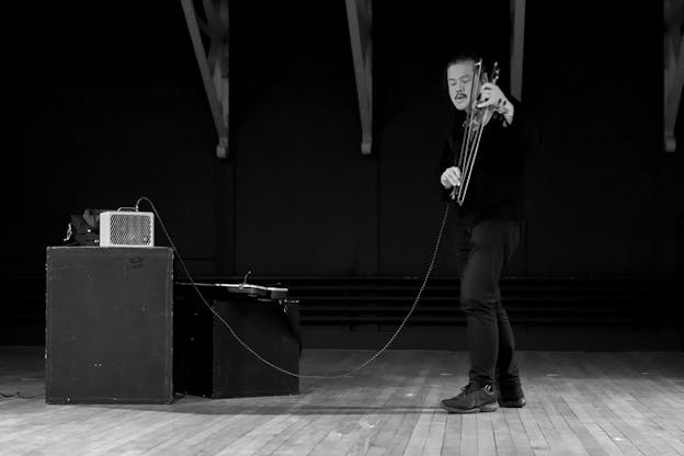 Black and white photograph of a person to the right playing a violing connected to wired leading to the left of the image on a speaker on top of a wooden box.