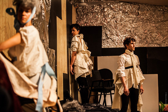 Three figures dressed in white sheet-like material clothing stand unmoving in a room whose walls are covered by aluminum foil. The one closest to the viewer kneels, as the other two furthest away keep their backs to each other.