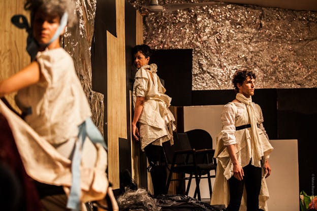 Three figures dressed in white sheet-like material clothing stand unmoving in a room whose walls are covered by aluminum foil. The one closest to the viewer kneels, as the other two furthest away keep their backs to each other.