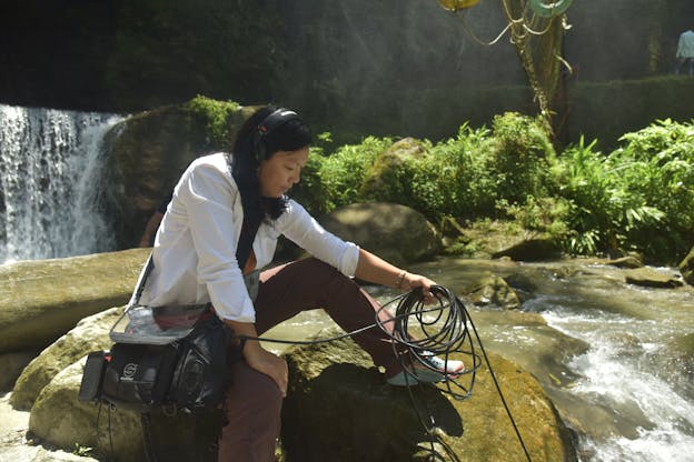A photograph of Susie Ibarra sitting on a rock in a body of water, wearing a white shirt, purple pants, headphones, and other recording equipment. She holds a cluster of black cables which extends into the water. Behind her, there is a small waterfall and a collection of light green bushes. 