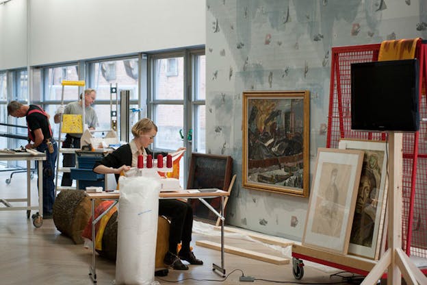 A person sits in front of a sewing machine holding up fabric while behind them one person works at a table and another carries a ladder, in a natural-lit room in which framed paintings are stacked on a red carrier against a gray wall. 