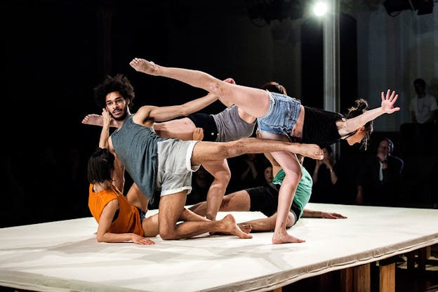 Three performers, two upright and one kneeling, extend their legs in arabesque while two performers weave their bodies through them on the floor.