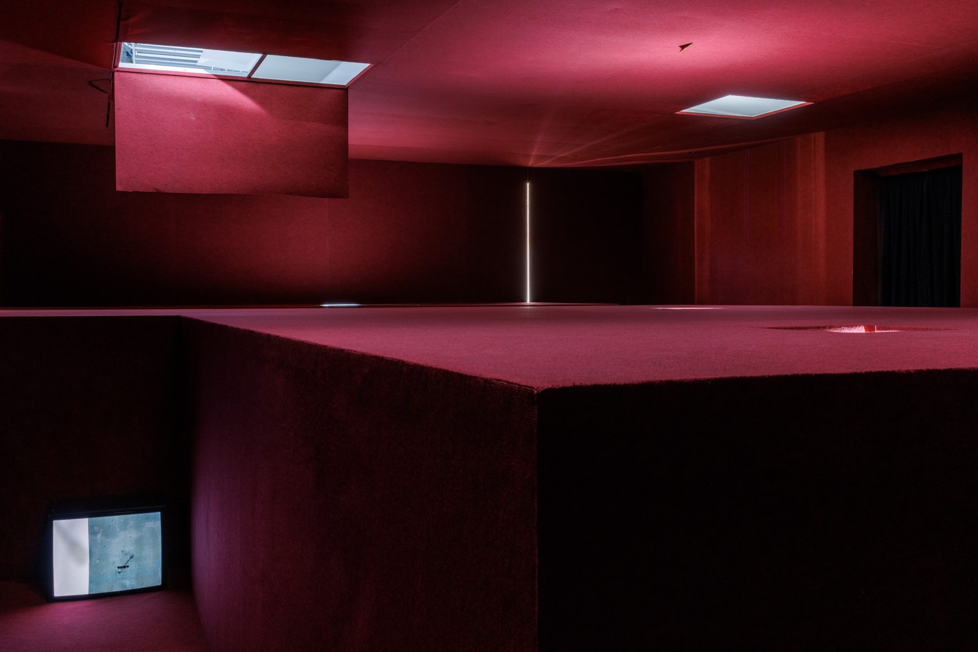 Installation by Pan Daijing. The labyrinthine space is covered entirely in red carpet-like material, except for a couple openings in the ceiling, through which soft light is visible. To the left is seen a tall recess which separates the different floors of the space. A television plays a video work by the artist, casting soft light onto the red floor and walls surrounding it.