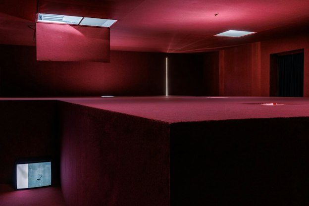 Installation by Pan Daijing. The labyrinthine space is covered entirely in red carpet-like material, except for a couple openings in the ceiling, through which soft light is visible. To the left is seen a tall recess which separates the different floors of the space. A television plays a video work by the artist, casting soft light onto the red floor and walls surrounding it.