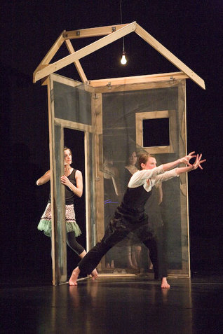 One performer wearing a black camisole over a white blouse and black pants is in a lunge position facing right in front of a simplified wooden house structure. They lift their arms in the front with fingers spread apart. Multiple performers stand behind the house structure and look towards the central performer. The performance is staged on a black floor against a black backdrop.