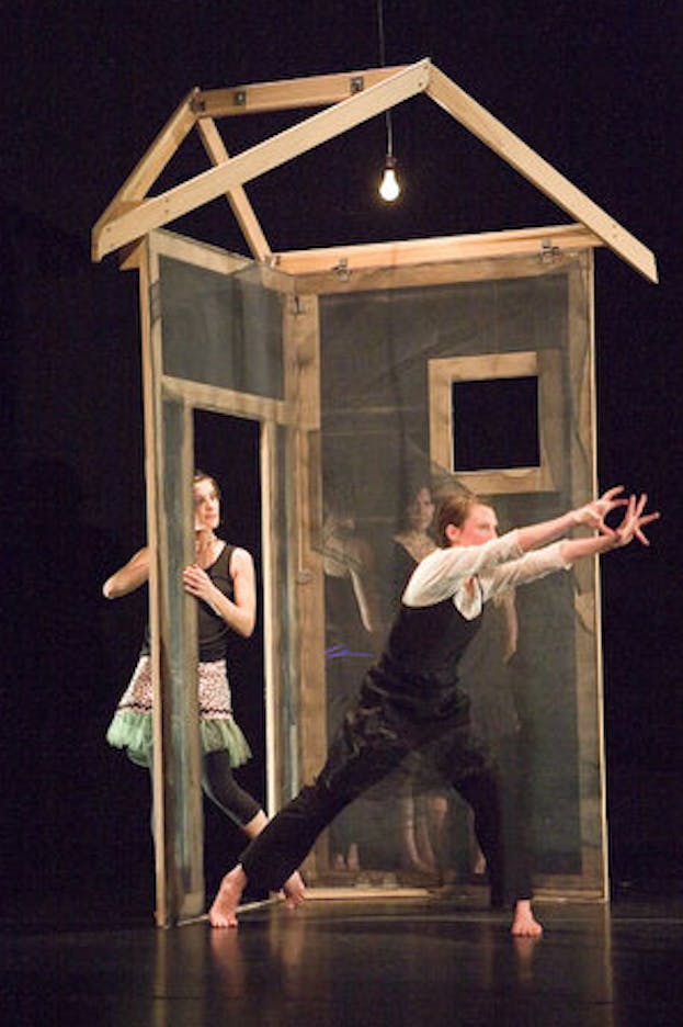 One performer wearing a black camisole over a white blouse and black pants is in a lunge position facing right in front of a simplified wooden house structure. They lift their arms in the front with fingers spread apart. Multiple performers stand behind the house structure and look towards the central performer. The performance is staged on a black floor against a black backdrop.