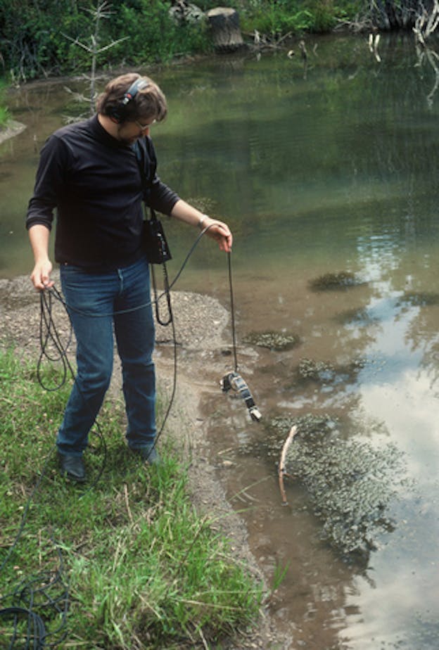 Dunn standing beside a murky green, algae-filled pond dangling a device attached to headphones above the water. 