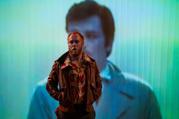 Dimly-lit performer wearing a leather jacket and red peony patterned shirt stands with their mouth slightly open in front of a cyan and turquoise projection of a blue-hued man's face and shoulders.